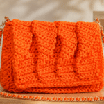 Bolsa de crochê laranja vibrante com textura em relevo e alça de corrente completa, exposta sobre uma superfície de madeira em ambiente minimalista e elegante.