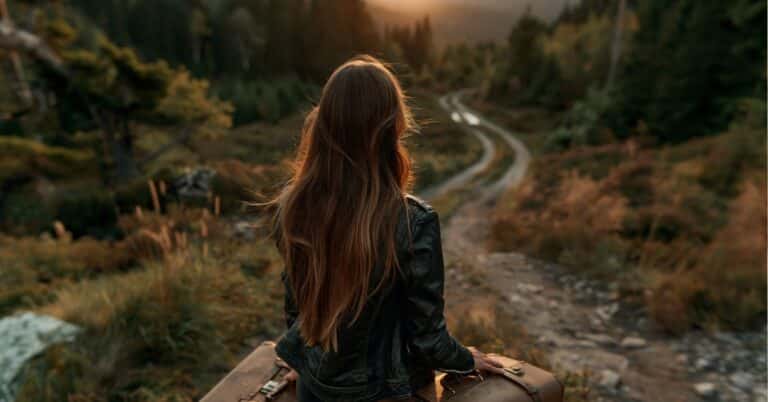 Mulher de costas usando jaqueta de couro, observando um caminho em meio à natureza ao pôr do sol.